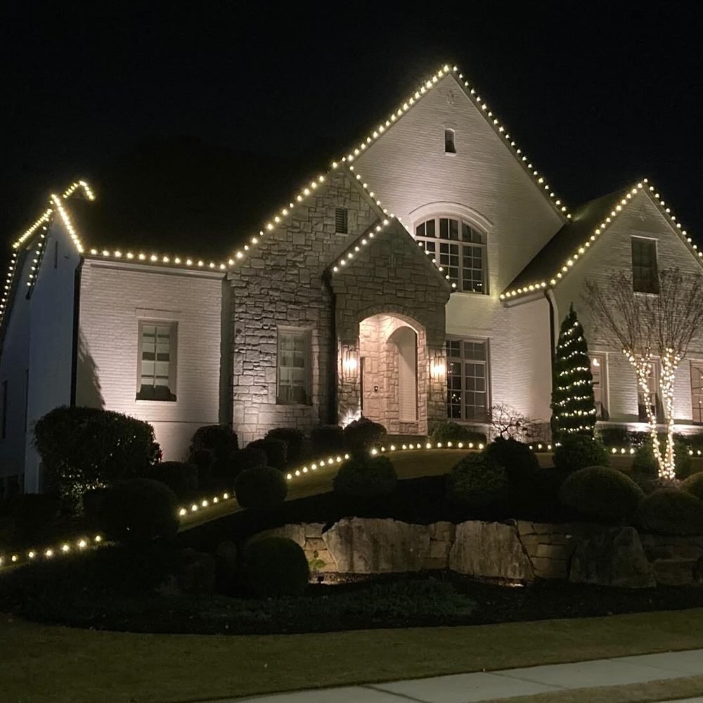 Stone and white-brick home at night, every gable run in warm white with a lit arched entry and cone trees